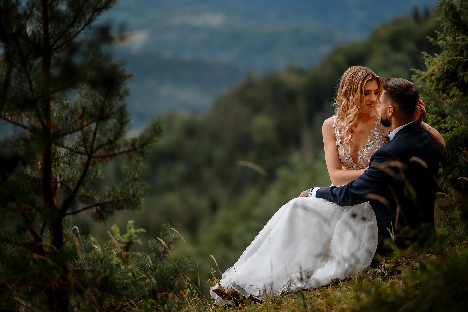Para młoda siedząca na wzgórzu z widokiem na zalesione góry, panna młoda w sukni z koronkową górą i długim spływającym dołem, pan młody w granatowym garniturze.