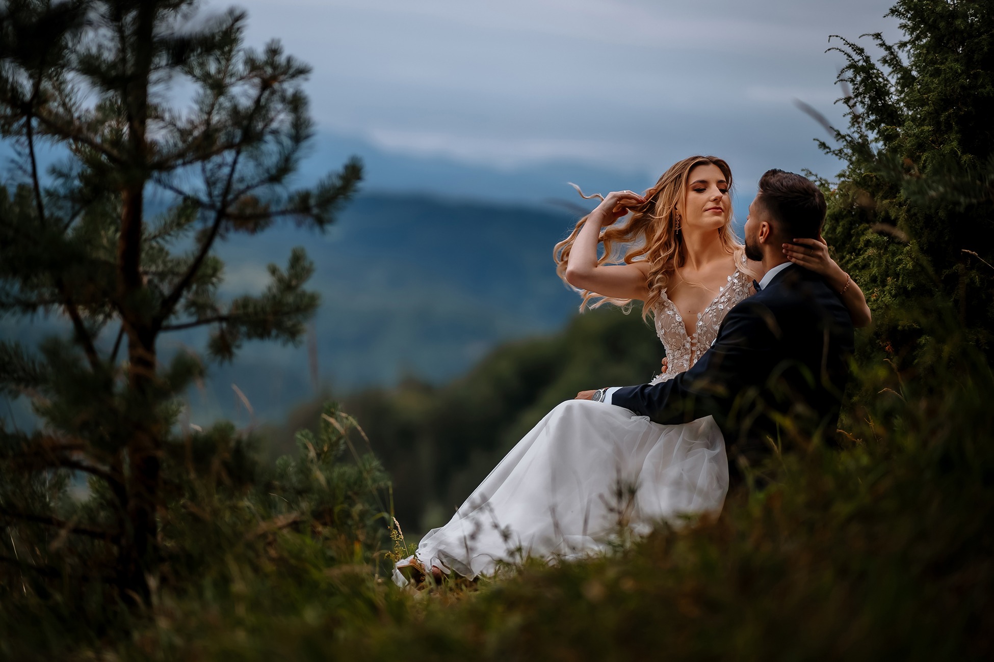 Para młoda siedząca na wzgórzu z widokiem na góry, panna młoda w sukni z koronkową górą, pan młody w ciemnym garniturze, romantyczny nastrój, naturalne otoczenie.