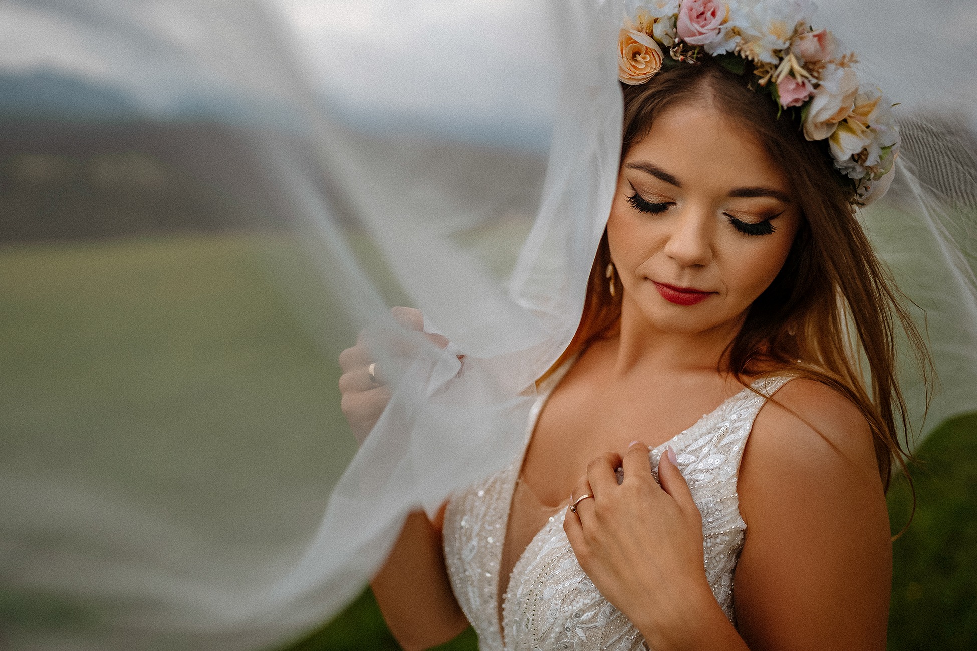 Panna młoda z wiankiem z jasnych kwiatów, trzymająca welon na tle rozmytego krajobrazu.