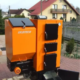 Pomarańczowo-czarny piec z podajnikiem tłokowym EKOTECH, umieszczony na palecie, z otwartymi drzwiczkami popielnika, widoczne elementy sterowania i wentylator
