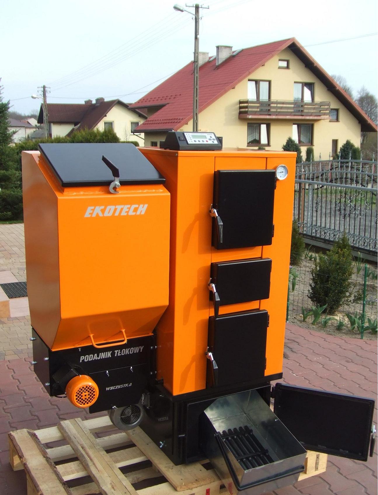 Pomarańczowo-czarny piec z podajnikiem tłokowym EKOTECH, umieszczony na palecie, z otwartymi drzwiczkami popielnika, widoczne elementy sterowania i wentylator