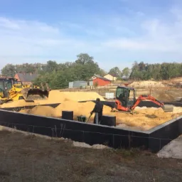 Wykop pod fundamenty domu jednorodzinnego, zabezpieczony czarną izolacją, wypełniony piaskiem; na terenie budowy żółta koparko-ładowarka i pomarańczowa mini koparka, pracownik z łopatą, w tle...