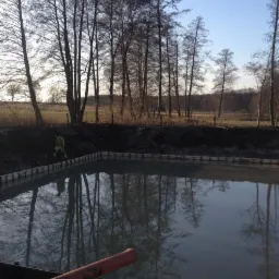 Wykopany staw z drewnianym szalunkiem brzegowym, wypełniony wodą, odbijający drzewa w tle. Widoczny fragment pomarańczowej rury i sylwetka pracownika w odblaskowej kamizelce.