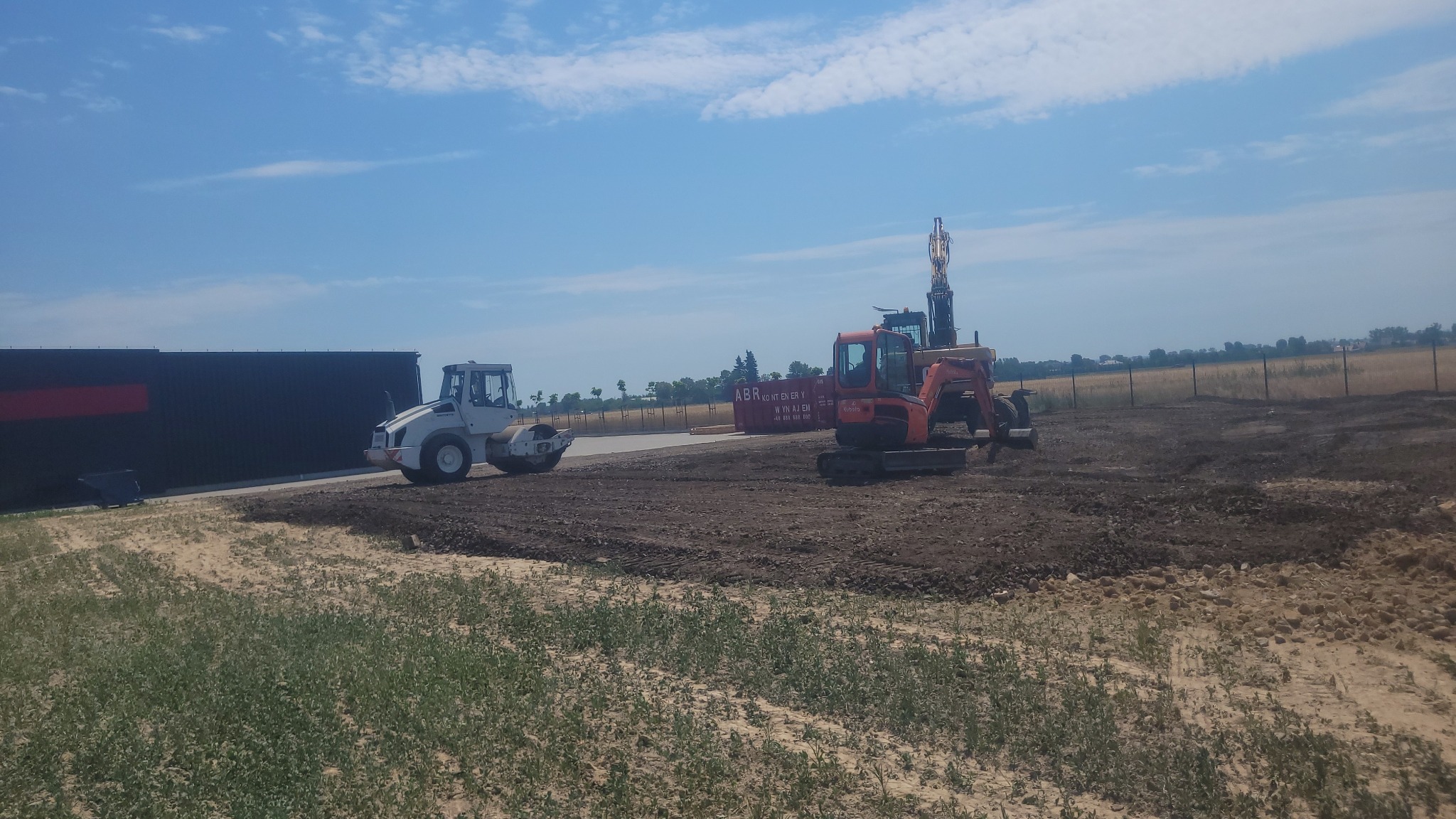 Niwelacja terenu w Parzynowie: walec drogowy i koparka na przygotowanym gruncie, kontener w tle. Błękitne niebo z delikatnymi chmurami.