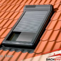 Okna dachowe z roletami zewnętrznymi, zamontowane na dachu pokrytym czerwoną dachówką ceramiczną, z widocznym logo producenta w prawym dolnym rogu.