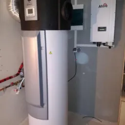 Biały zasobnik ciepłej wody użytkowej marki Sofath z czarną górną częścią i srebrnym przewodem wentylacyjnym, obok biała skrzynka inwertera Afore na szarej ścianie w surowym pomieszczeniu.