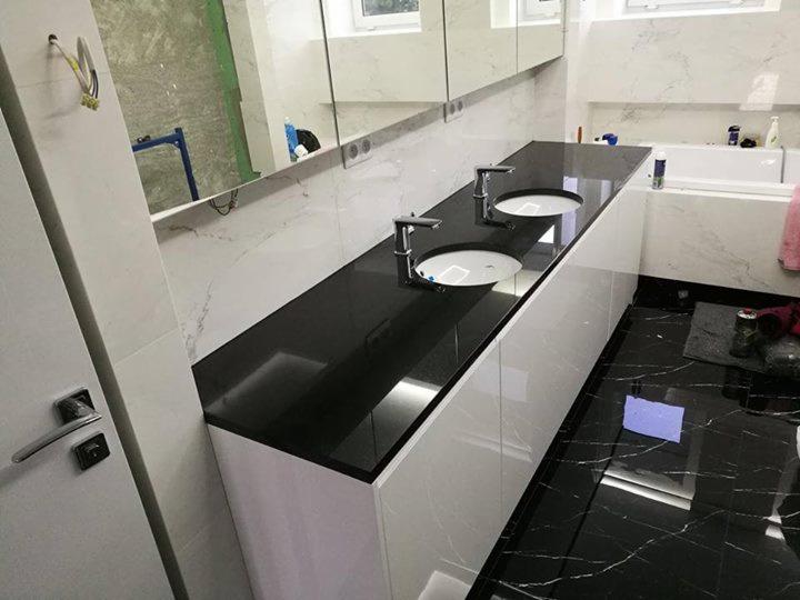 Czarny blat kamienny z dwiema umywalkami w łazience wykończonej białym marmurem i czarną marmurową podłogą, widoczne niedokończone prace instalacyjne.