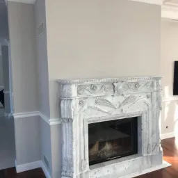 Elegancki marmurowy portal z paleniskiem, zdobiony roślinnymi motywami, w jasnym wnętrzu z drewnianą podłogą i dekoracyjnymi listwami przysufitowymi.