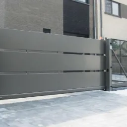 Automatyczna, przesuwna brama w kolorze grafitowym z poziomymi prześwitami, zamontowana na posesji z ogrodzeniem panelowym, widoczne budynki w tle.