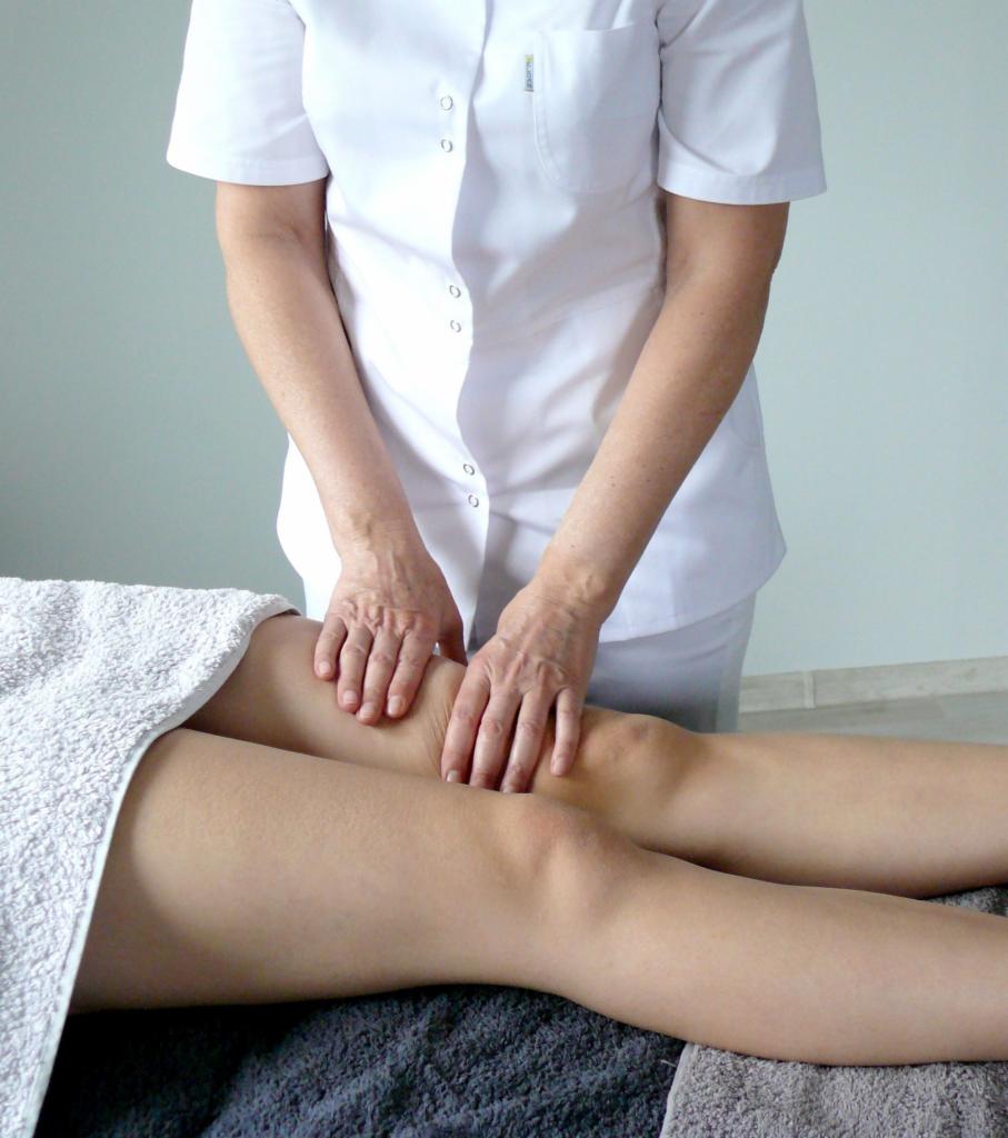 Terapeuta w białym uniformie wykonuje drenaż limfatyczny na nogach pacjenta leżącego na stole przykrytym ręcznikiem.