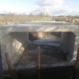 Gotowy betonowy przepust skrzynkowy na rzece, widoczne uchwyty na górnej powierzchni, drewniane pale w wodzie i ziemne brzegi rzeki.