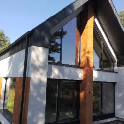 Nowoczesny dom z białym tynkiem, dużymi oknami z czarnymi ramami i grafitowym dachem, otoczony sosnowym lasem w słoneczny dzień.