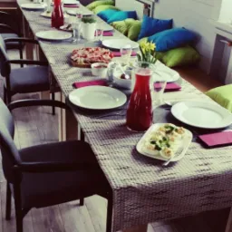 Stół nakryty do przyjęcia z przekąskami, napojami w dzbankach i dekoracjami kwiatowymi. Widoczne krzesła i poduszki do siedzenia w tle.