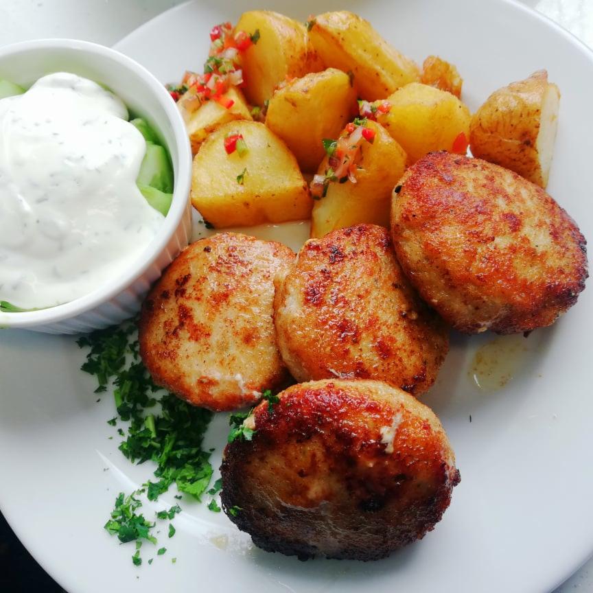 Apetyczne kotleciki z ziemniakami posypanymi kolorową salsą i sosem tzatziki w białej miseczce, całość udekorowana świeżą natką pietruszki na białym talerzu.