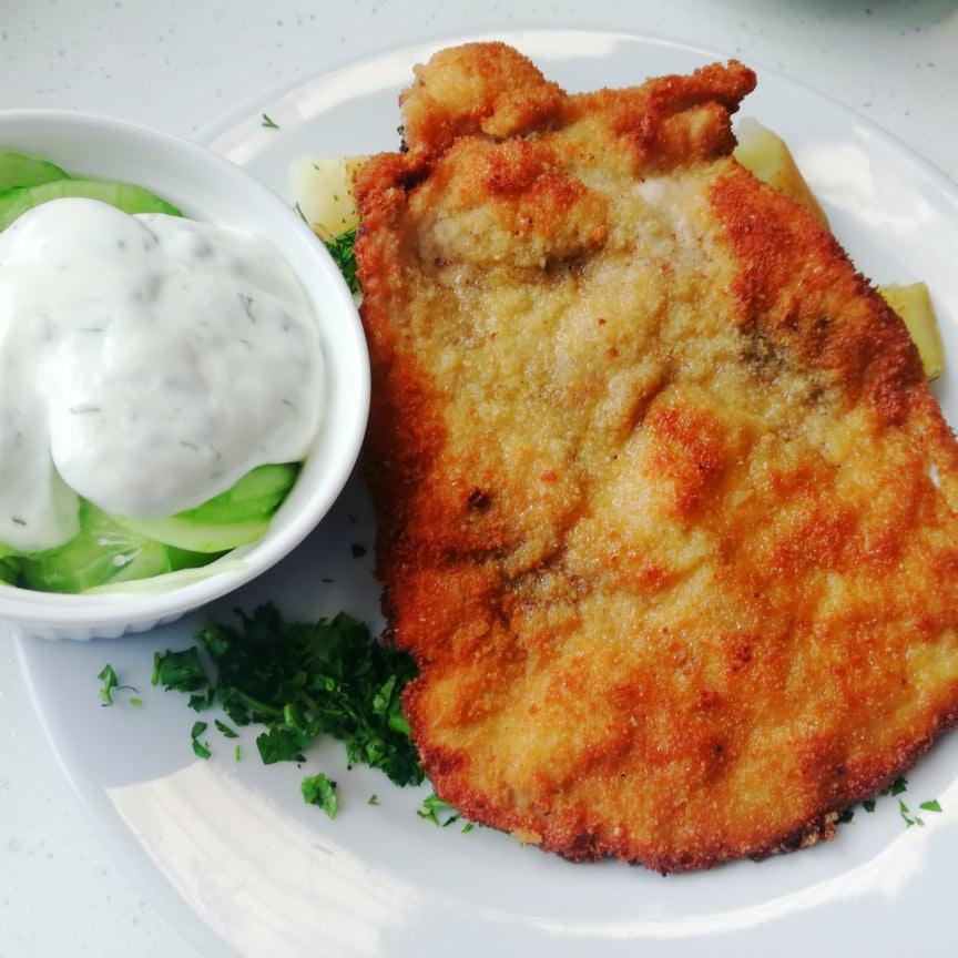 Smażony sznycel w złocistej panierce podany z ogórkiem w śmietanie i posiekaną pietruszką na białym talerzu.