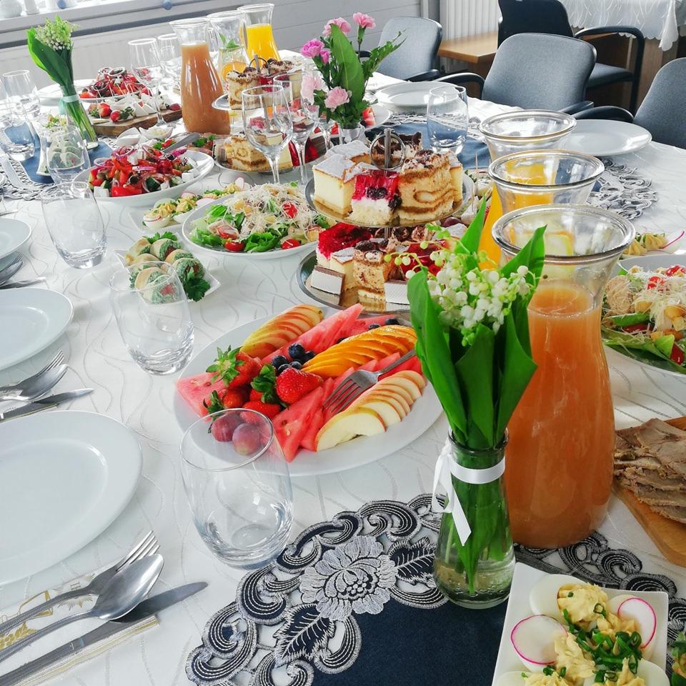Uroczysty stół z białym obrusem, zastawiony owocami, ciastami, sałatkami i dzbankami z sokiem, udekorowany konwaliami w wazonach.