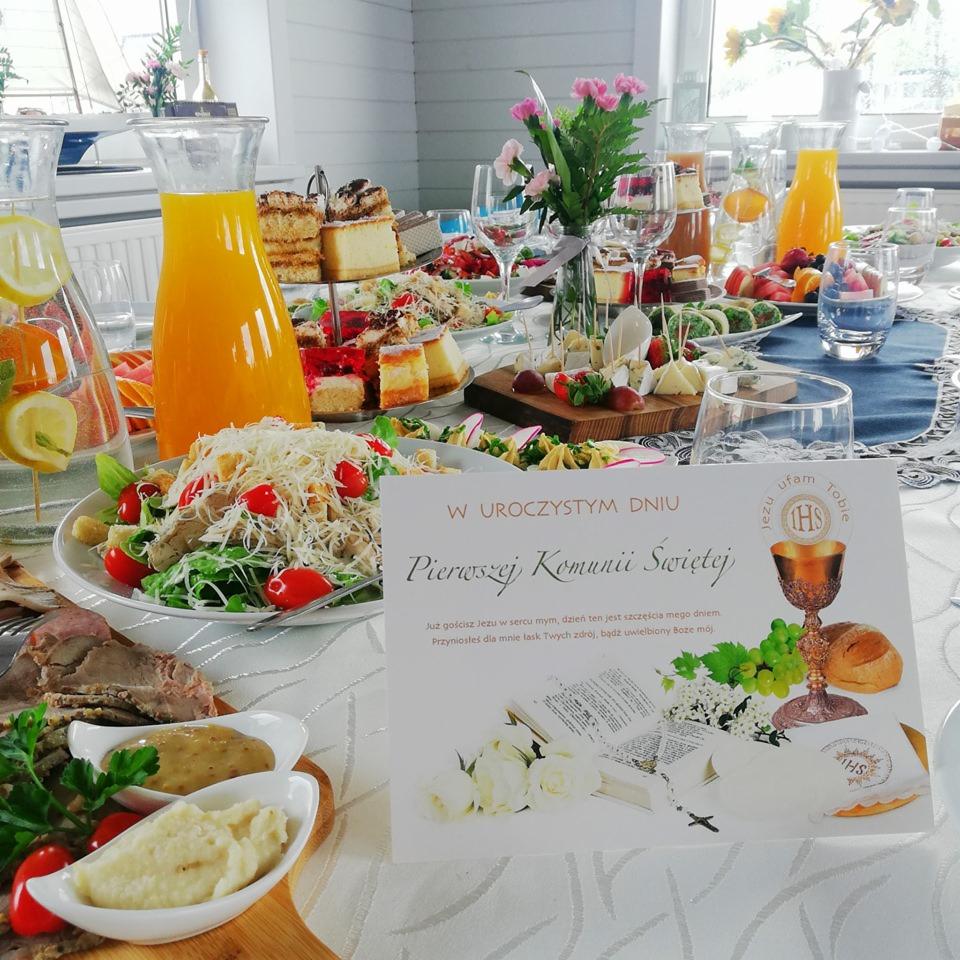 Bogato zastawiony stół z potrawami i napojami, przygotowany na uroczystość Pierwszej Komunii Świętej w Zegrzu Południowym, z widoczną kartką okolicznościową i dekoracjami kwiatowymi.