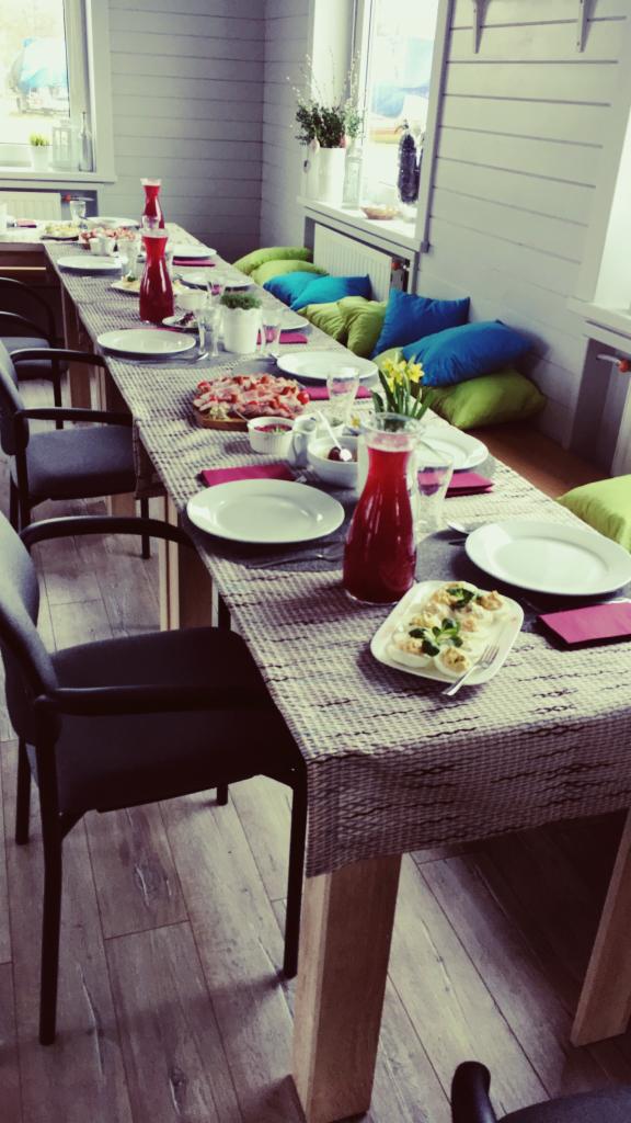 Stół nakryty do przyjęcia z przekąskami, napojami w dzbankach i dekoracjami kwiatowymi. Widoczne krzesła i poduszki do siedzenia w tle.