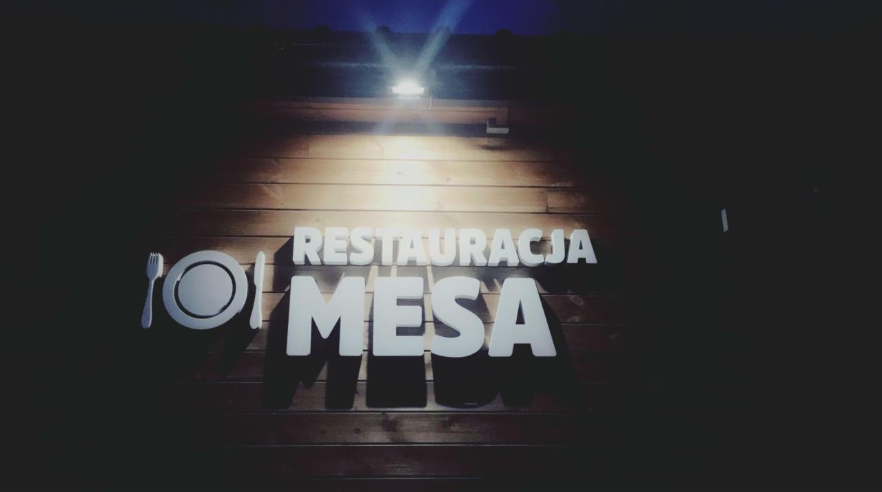 Podświetlony szyld restauracji 'MESA' z logo talerza, widelca i noża na drewnianej ścianie, oświetlony od góry.