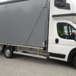 Biały samochód dostawczy z szarą plandeką, widok z boku na szarym brukowanym podłożu, widoczny napis 'DAR' na plandece.