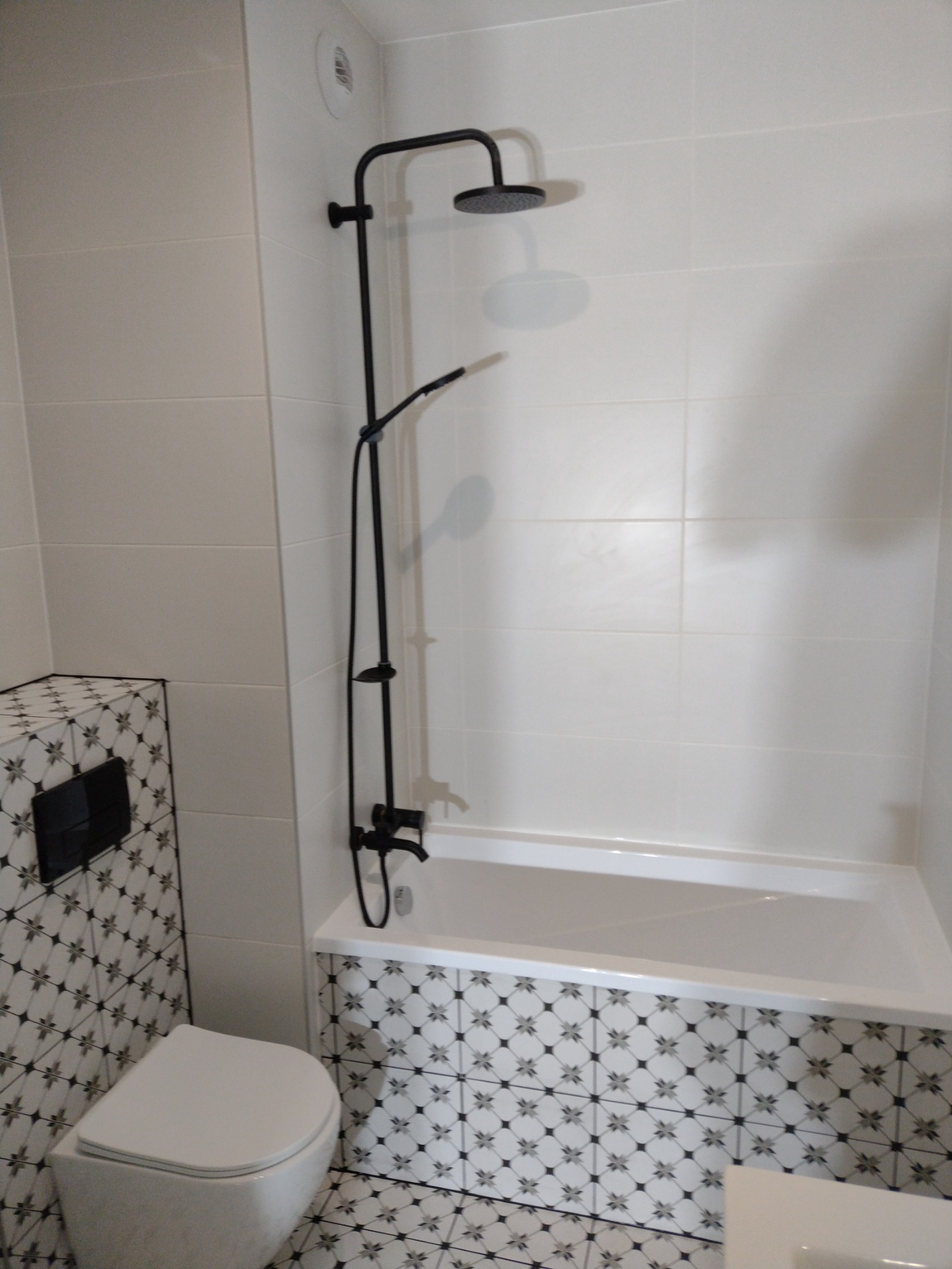 Nowoczesna łazienka w Pabianicach z białą wanną, czarną armaturą prysznicową i wzorzystymi płytkami ceramicznymi na obudowie wanny oraz podłodze.