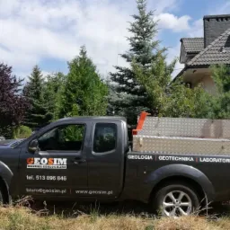 Szara terenowa ciężarówka z logo firmy Geosim i sprzętem do wierceń geologicznych na pace, zaparkowana na trawie przed domem.