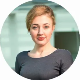 Portret kobiety z blond włosami i szarym topem na rozmytym tle biurowym.