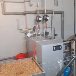 Szary piec na pellet z zasobnikiem pełnym pelletu, widoczne rury instalacji grzewczej, manometr i pompy obiegowe.