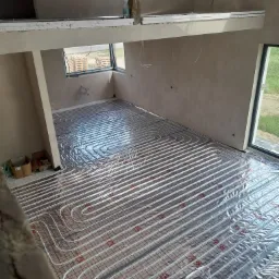 Białe rury ogrzewania podłogowego ułożone spiralnie na folii z czerwoną siatką w surowym wnętrzu z oknami.