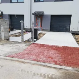 Nowo ułożona, czerwona kostka brukowa w jodełkę przed budynkiem z szarą stolarką, widoczny fragment traktora i świeżo ułożone, szare płyty chodnikowe.