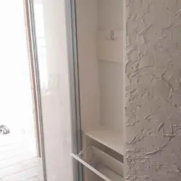 Biała szafa wnękowa z przesuwnymi drzwiami i wysuwanymi półkami na buty, widoczne wieszaki na ubrania, ściana z fakturą.