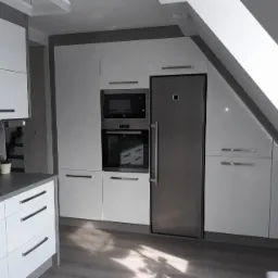 Biała kuchnia z zabudową pod skosem, srebrna lodówka i piekarnik w zabudowie, minimalistyczne uchwyty, szare blaty i podłoga.