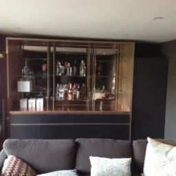 Domowy bar na wymiar z przeszkloną witryną, podświetlony, z zapasem alkoholu, widok z perspektywy sofy w salonie.