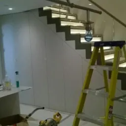 Montaż szafy wnękowej pod schodami z oświetleniem LED, widoczna drabina i narzędzia pracy.