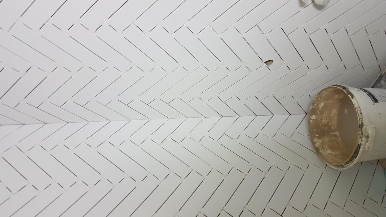 Ułożona biała glazura w jodełkę na ścianie, widoczny wiadro z zaprawą i niedokończone fugowanie.