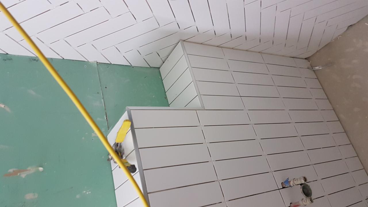 Układanie białych, podłużnych płytek na ścianach i suficie łazienki, widoczna zielona płyta gipsowo-kartonowa i narzędzie do cięcia płytek z żółtą rączką.