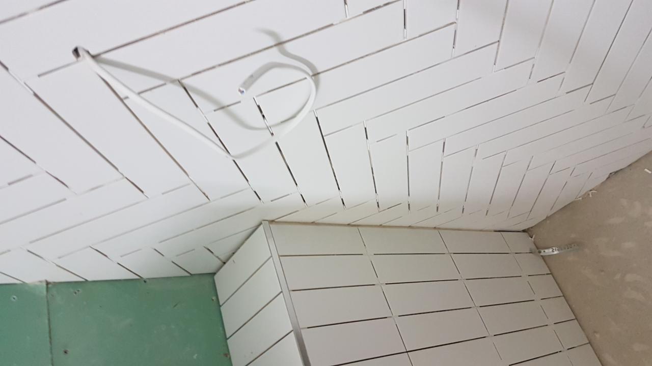 Białe, podłużne płytki ułożone w jodełkę na ścianie i suficie łazienki, widoczny przewód elektryczny i fragment zielonej płyty gipsowej.