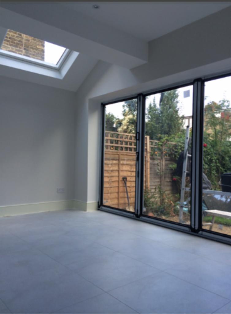 Wnętrze nowocześnie wykończonego pokoju z szarymi płytkami podłogowymi i składanymi, przeszklonymi drzwiami balkonowymi wychodzącymi na ogród. Widoczny fragment drabiny oparty o ogrodzenie.