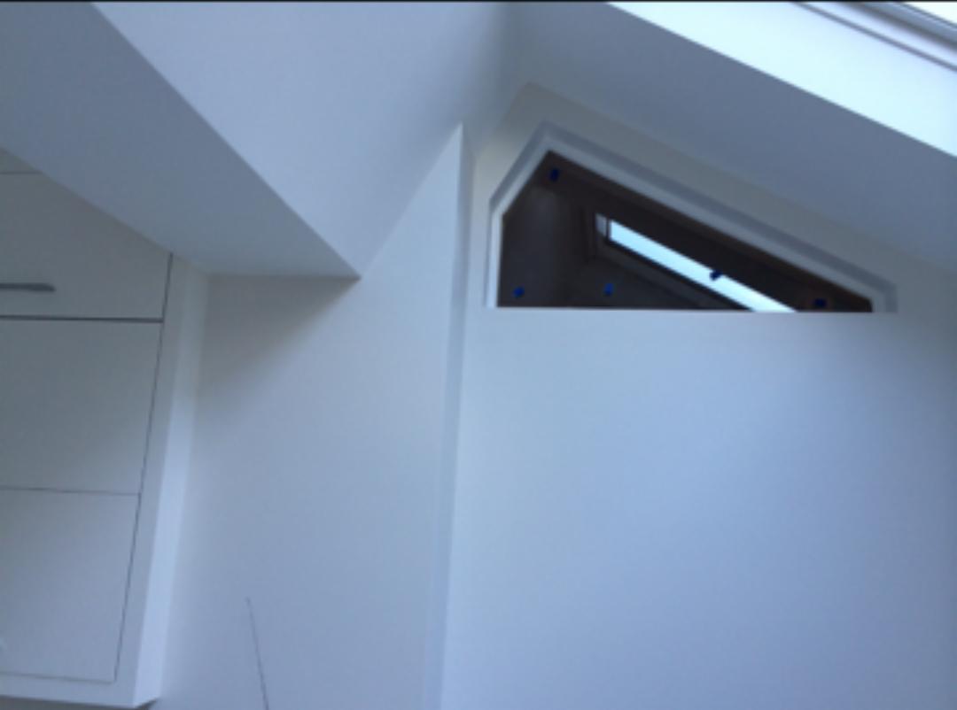 Świeżo pomalowane, białe wnętrze poddasza z oknem dachowym w kształcie trapezu, zabezpieczonym niebieską taśmą malarską, z fragmentem zabudowy kuchennej po lewej stronie.