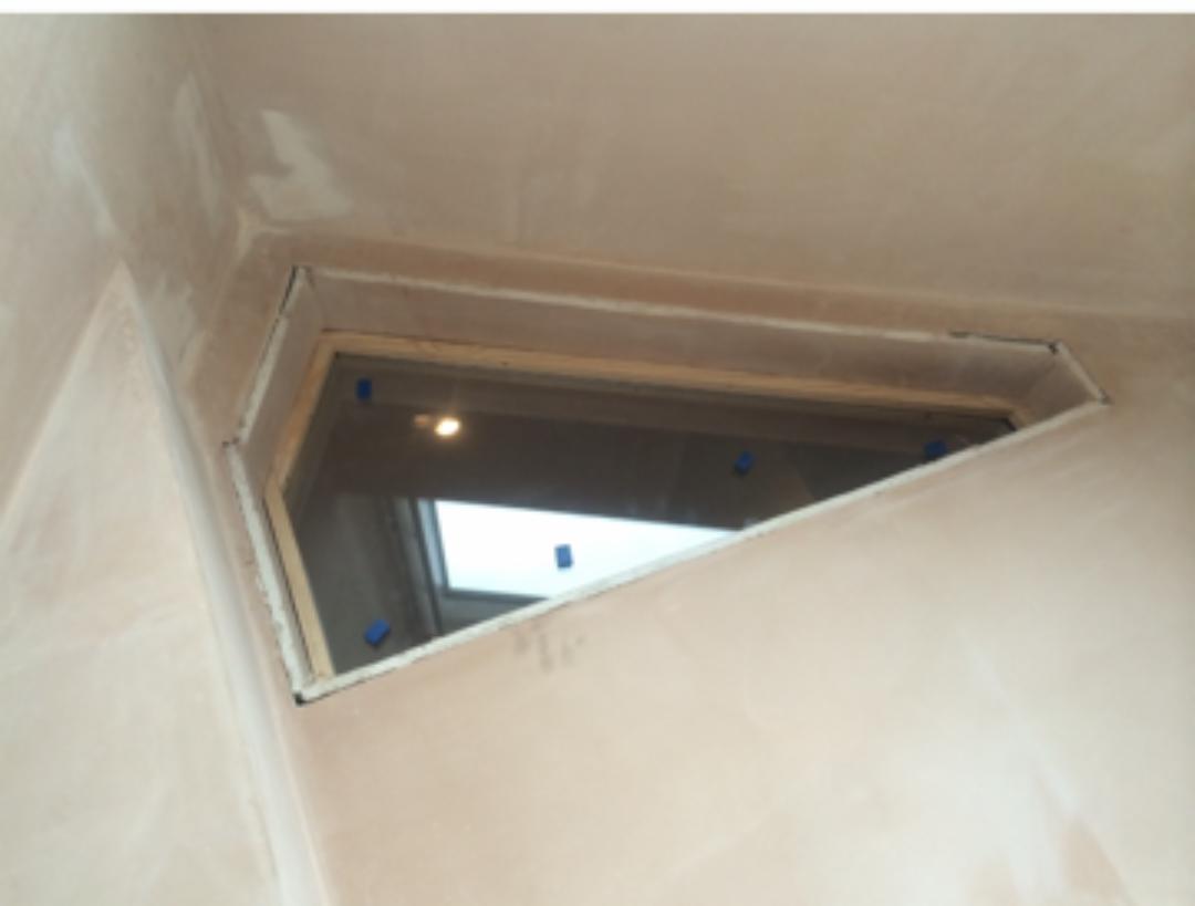 Nietypowe okno w kształcie trapezu w ścianie po świeżym tynkowaniu, zabezpieczone niebieską taśmą malarską.