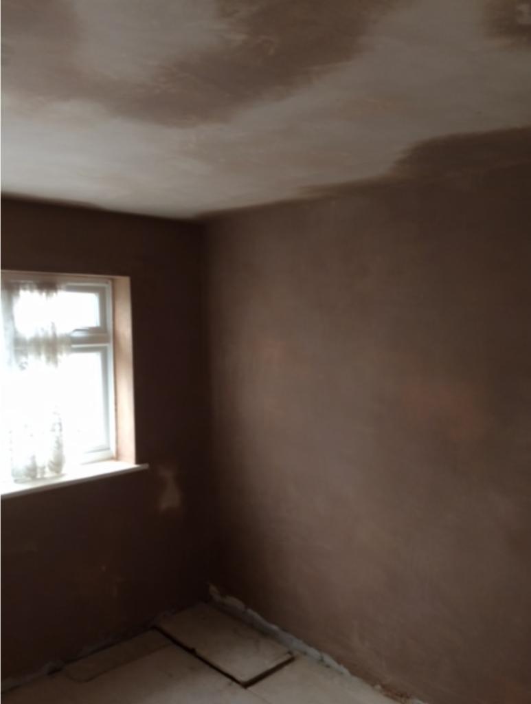 Pokój po nałożeniu nowej warstwy tynku na ścianach i suficie, widoczne niedoskonałości na suficie, okno z firanką w lewym górnym rogu, podłoga częściowo zakryta płytami.