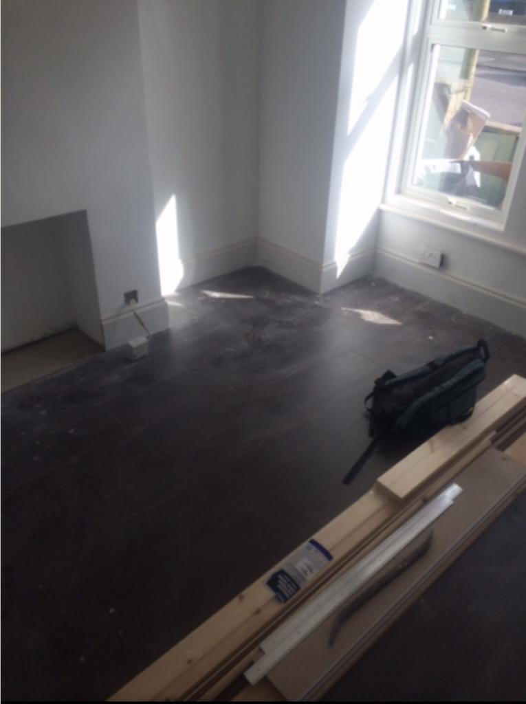 Pokój w trakcie prac wykończeniowych: widoczne białe ściany, ciemna podłoga, listwy przypodłogowe, okno z silnym światłem słonecznym, torba robocza i stos drewnianych desek.