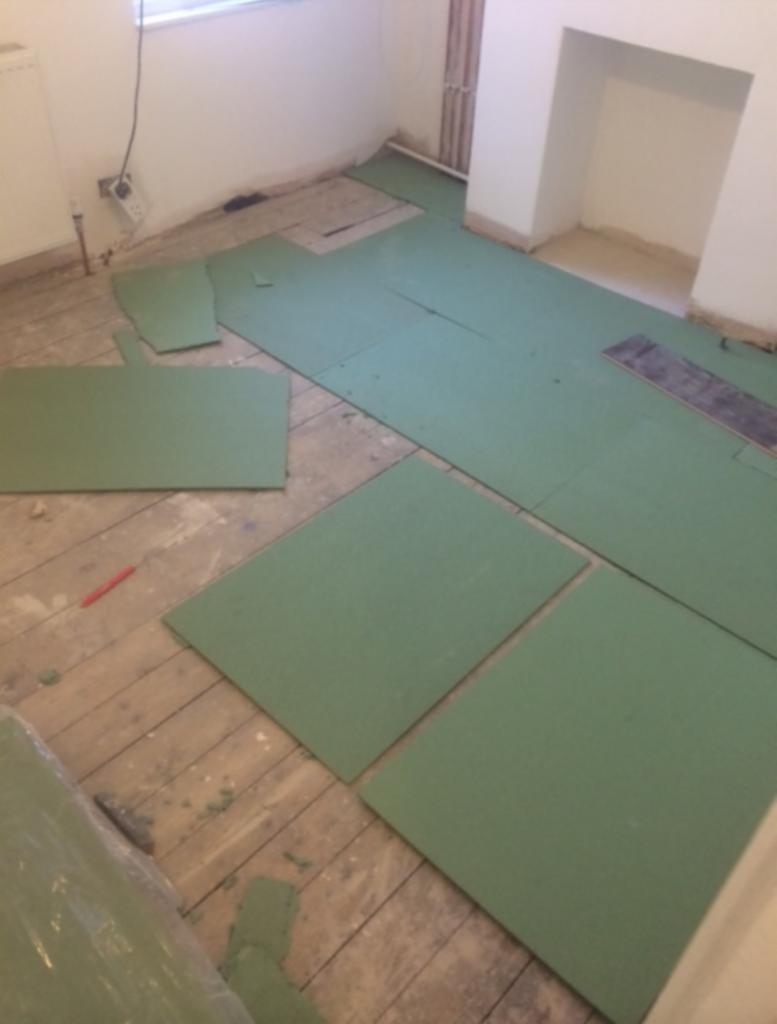 Przygotowanie podłoża w łazience: zielone płyty izolacyjne rozłożone na starej drewnianej podłodze, widoczny fragment rolki z materiałem izolacyjnym i czerwony ołówek.