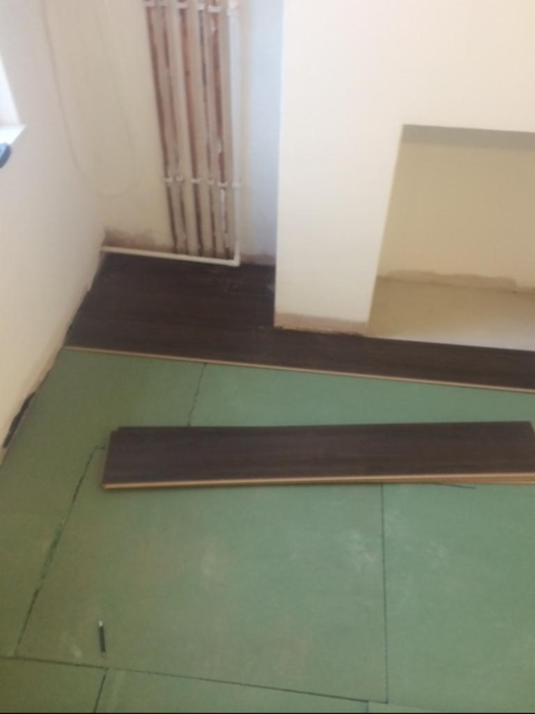 Podłoga z paneli w trakcie układania, widoczna zielona mata podkładowa, częściowo ułożone ciemne panele, grzejnik ścienny i fragment niezabezpieczonej ściany.