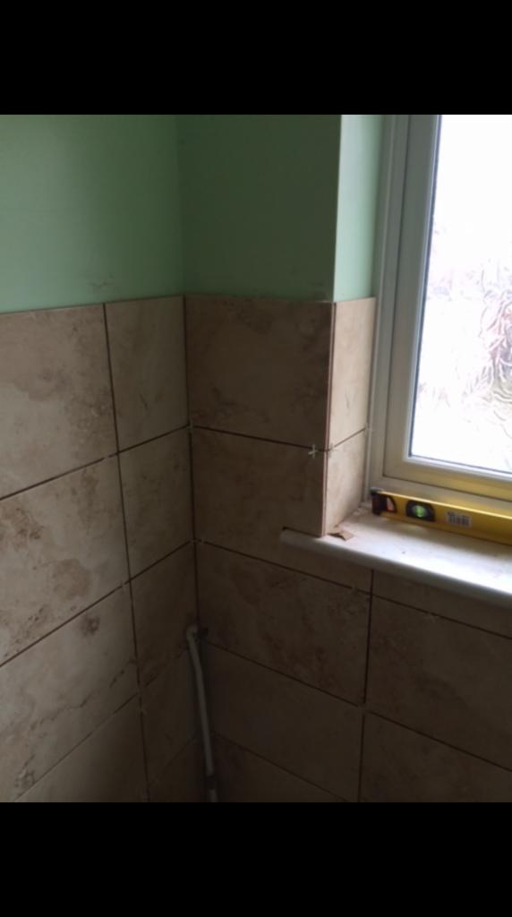 Układanie beżowych płytek imitujących marmur w narożniku łazienki, widoczne krzyżyki dystansowe i poziomnica na parapecie okna.