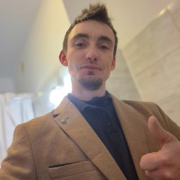 DAMPLEKS DAMIAN SZAJERSKI - Mężczyzna w brązowej marynarce, z uniesionym kciukiem do góry, robiący selfie w jasnym wnętrzu. Widoczny fragment ściany z teksturą.