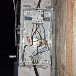 DAMPLEKS DAMIAN SZAJERSKI - Otwarta skrzynka elektryczna z widocznymi przewodami i bezpiecznikiem, zamontowana na szarej ścianie z betonu komórkowego, obok drewnianej ściany. Widoczne złącza i kable.