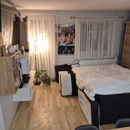 DAMPLEKS DAMIAN SZAJERSKI - Jasne wnętrze sypialni z łóżkiem, drewnianą podłogą i szafą. Dekoracyjne oświetlenie i plakat na ścianie dodają przytulności. Widok na uporządkowane pomieszczenie.