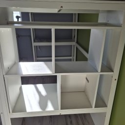 DAMPLEKS DAMIAN SZAJERSKI - Biały regał z półkami, widok z dołu, ukazujący konstrukcję i mocowania. Widoczne elementy instalacji w tle. Ujęcie z perspektywy podłogi, podkreślające montaż.