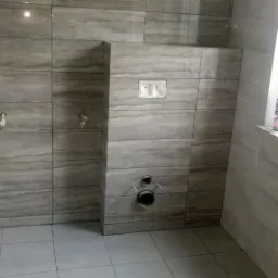 Świeżo wykafelkowana ściana i podłoga w łazience w odcieniach szarości, z widocznymi przyłączami hydraulicznymi i puszką podtynkową do spłuczki WC, jeszcze bez armatury.